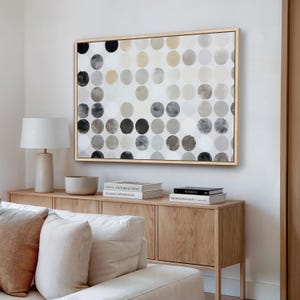 May include: Abstract wall art featuring a pattern of overlapping circles in shades of gray, black, and beige. The artwork is framed in a light wood frame and hangs above a wooden cabinet. The room also includes a lamp, books, and a sofa.