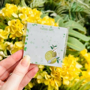 May include: A square Matcha Cafe notepad with a green and white design. The notepad features a cartoon illustration of a matcha roll cake with a spoon and a cat. The notepad is held in front of a background of yellow flowers.
