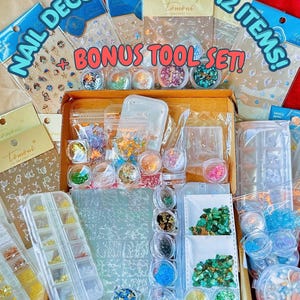 Könnte beinhalten: Eine offene Box voller Nailart-Zubehör, darunter Glitzer, Edelsteine und Aufkleber. Die Box enthält verschiedene Behälter mit farbenfrohen Nageldekorationen, mit dem Text "NAIL DECO SCOOP 10-12 ITEMS! + BONUS TOOL SET!"