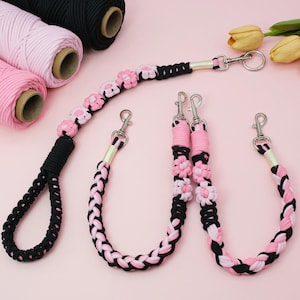 Op de afbeelding: Een collectie handgemaakte macramé hondenriemen en accessoires in zwart, roze en wit. De riemen hebben gevlochten ontwerpen en bloemaccenten, met metalen sluitingen. Spoelen met bijpassend macramé koord zijn ook zichtbaar.