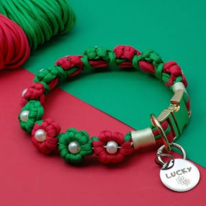 Puede incluir: Un collar de perro trenzado rojo y verde con un diseño de flores y una etiqueta plateada que dice "LUCKY" con un pequeño símbolo de flor.