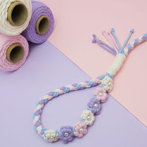 May include: A macrame cord accessory featuring a braided design in pastel pink, lavender, and cream colors. The cord is adorned with small fabric flowers, each centered with a white faux pearl bead. Several loose cord ends extend from one side.