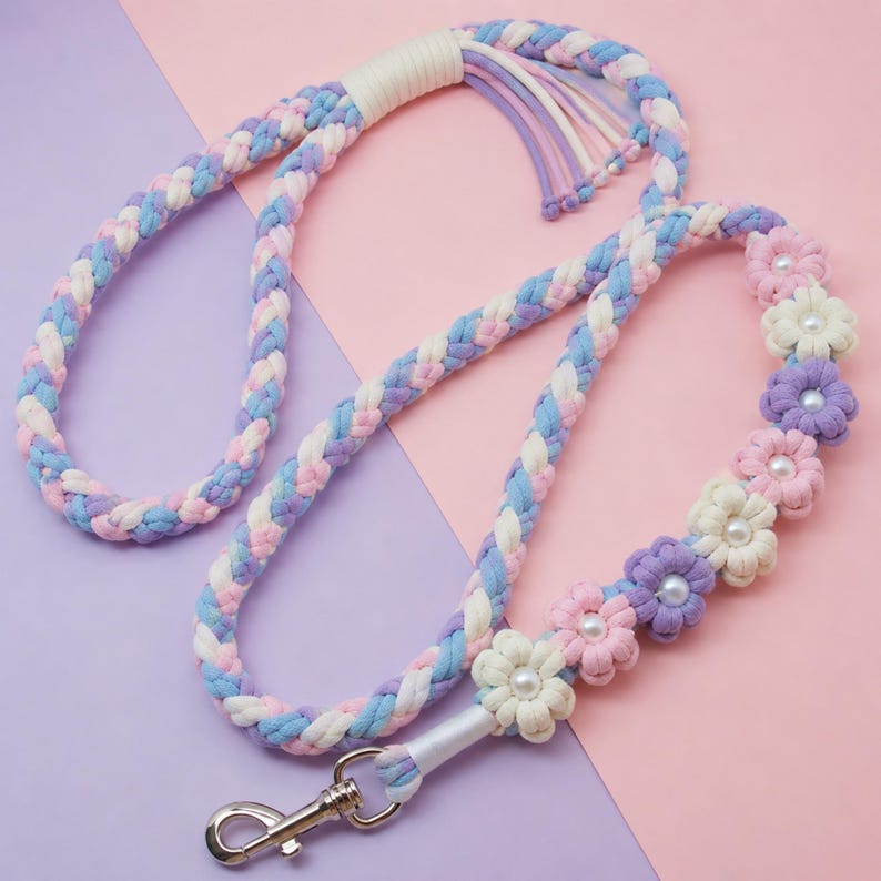 May include: A braided rope leash featuring a pastel color palette of light blue, soft pink, and lavender. The leash is accented with several fabric flowers in white, pink, and purple, each centered with a small white pearl bead. A silver metal swivel clasp is attached to one end.