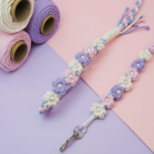 May include: Two macrame lanyards featuring a repeating floral pattern made from cream, light pink, and lavender cotton cord. Each flower center is accented with a white faux pearl bead. One lanyard includes a silver-toned metal swivel lobster clasp.