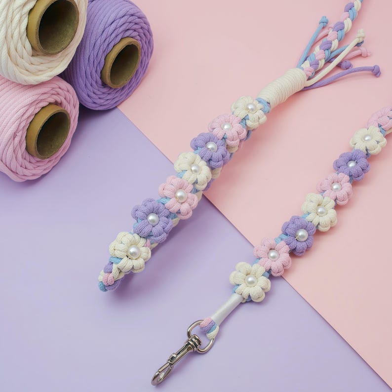 May include: Two macrame lanyards featuring a repeating floral pattern made from cream, light pink, and lavender cotton cord. Each flower center is accented with a white faux pearl bead. One lanyard includes a silver-toned metal swivel lobster clasp.