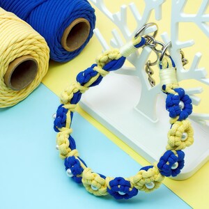 May include: A handmade blue and yellow macrame dog collar with pearl accents. The collar is displayed on a white decorative stand, with spools of blue and yellow macrame cord in the background. The collar is made of knotted rope.