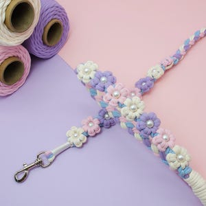 May include: A braided macrame lanyard featuring pastel pink, lavender, and light blue cord woven into flower shapes with white faux pearl centers. A silver-toned metal swivel lobster clasp is attached to one end of the floral cord strap.