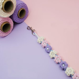 May include: A lanyard featuring a series of small, knotted fabric flowers in cream, light pink, and purple, each with a central white faux pearl bead. The floral strand is attached to a silver-toned metal swivel lobster clasp at the top end.