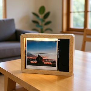 Op de afbeelding: Een houten fotodisplay met een lichte houten afwerking. Het bevat een stapel foto's, verlicht door een lichtstrip. Aan de zijkant bevindt zich een zwart paneel. De foto's tonen een stel tegen een zonsondergang. Het apparaat staat op een houten tafel.