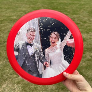 Puede incluir: Un frisbee rojo con una foto de una novia y un novio caminando lejos de una ceremonia. La foto muestra a la pareja sonriendo y siendo rociada con confeti.