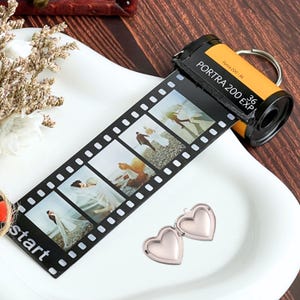 Pode incluir: Um chaveiro amarelo e preto em forma de rolo de filme com uma tira de fotos em preto e branco anexada. A tira de fotos apresenta imagens de um casal em um cenário de casamento. O chaveiro também inclui dois pingentes em forma de coração prateados.
