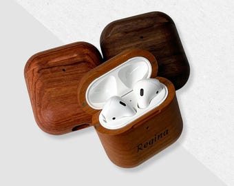 May include: A wooden case for wireless earbuds with a personalized inscription that reads "Regina". The case is open and the white earbuds are visible inside.