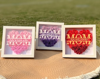 Può includere: Tre cornici bianche con un'arrangiamento a forma di cuore di rose di carta rosa, viola e rosse. Ogni cornice ha la parola "MOM" in lettere dorate con i nomi "Aiden, Ayvah e Alex" scritti sotto.