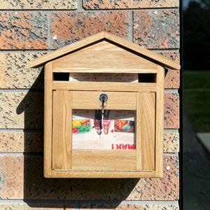 May include: A wooden mailbox with a pitched roof, mounted on a brick wall. The mailbox has a key lock and a clear window showing the contents. The word "MAKI" is visible on the newspaper inside.
