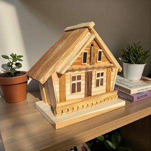 Puede incluir: Una maqueta de casa de madera hecha a mano con tejado a dos aguas, ventanas y chimenea. La casa est&aacute; hecha de tonos de madera claros y oscuros y se asienta sobre una base rectangular. El modelo se exhibe en una estanter&iacute;a de madera.