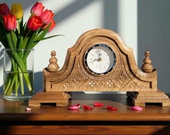 Puede incluir: Un reloj de sobremesa de madera con esfera blanca y números romanos. El reloj tiene tallas ornamentadas y está flanqueado por pináculos decorativos. Un jarrón de tulipanes rojos y amarillos está a la izquierda.