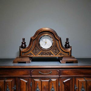 May include: A wooden mantel clock with a light-colored clock face and ornate carvings. The clock has a dark brown finish and decorative finials on each side. The clock is sitting on a dark surface, and the background is a solid gray.