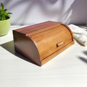 May include: A wooden bread box with a roll-top lid and a small handle. The box is made of light brown wood and has a curved front. The bread box is sitting on a white surface.