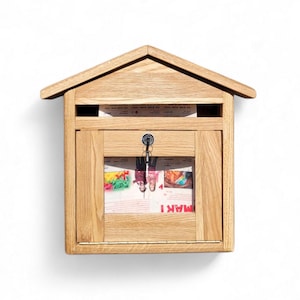 May include: A wooden mailbox with a house-shaped top. The mailbox is made of light-colored wood and has a door with a key lock. Inside the door is a magazine with the word "MAKI" visible. The mailbox is designed to be mounted on a wall.