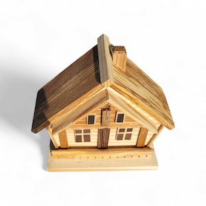 Puede incluir: Una peque&ntilde;a casa de madera con tejado a dos aguas y ventanas. La casa est&aacute; hecha de madera marr&oacute;n clara y oscura, con un dise&ntilde;o detallado. La casa se asienta sobre una base de madera con una valla decorativa. La casa mide unos 15 cm de largo.