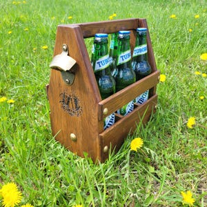 Könnte beinhalten: Ein Bierkasten aus Holz mit eingebautem Flaschenöffner, der vier grüne Glasbierflaschen enthält. Der Kasten ist braun mit Messingnieten und einem Griff. Die Flaschen haben blaue Verschlüsse und das Wort "TUBOR".