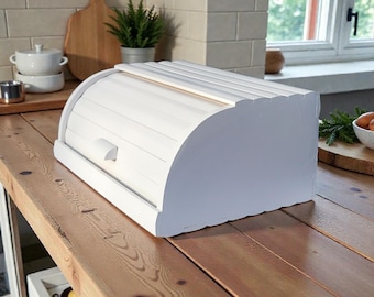 Può includere: Una cassetta per il pane bianca con apertura a rullo e design curvo è appoggiata su un piano di lavoro in legno. La cassetta ha un top a doghe e una piccola maniglia. Un tagliere, una piccola pianta e altri oggetti da cucina sono sullo sfondo.