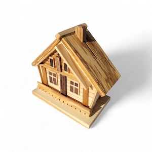 Puede incluir: Una hucha de madera con forma de casa con tejado a dos aguas, chimenea y peque&ntilde;as ventanas. La hucha est&aacute; construida con madera clara con vetas visibles, lo que le da un aspecto r&uacute;stico. El dise&ntilde;o incluye una peque&ntilde;a estructura similar a un porche en la base.