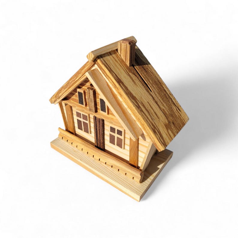 Puede incluir: Una hucha de madera con forma de casa con tejado a dos aguas, chimenea y peque&ntilde;as ventanas. La hucha est&aacute; construida con madera clara con vetas visibles, lo que le da un aspecto r&uacute;stico. El dise&ntilde;o incluye una peque&ntilde;a estructura similar a un porche en la base.