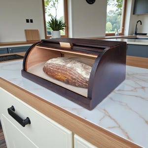 May include: A dark brown wooden bread box with a roll-top lid, showcasing a loaf of bread inside. The box has a curved design and is placed on a white countertop with a marble pattern. The bread is on a sheet of parchment paper.