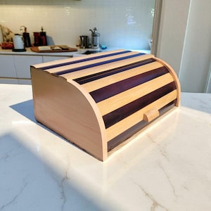 May include: Wooden bread bin with a curved top and a light wood finish. The top has alternating dark and light wood stripes. A small wooden handle is on the front. The bread bin is on a white countertop.