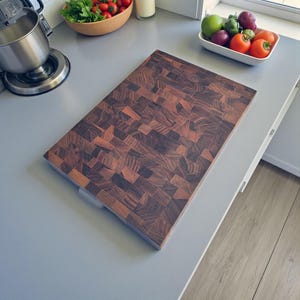 Puede incluir: Una tabla de cortar rectangular de extremo de grano con un patrón de mosaico marrón oscuro y rojizo. La tabla tiene un asa de color claro en un lado. La tabla está sobre una encimera gris.