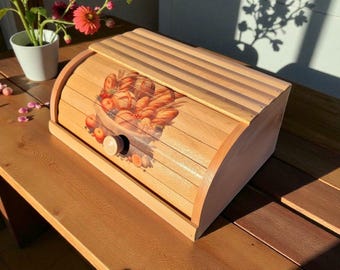 May include: A wooden bread box with a roll-top lid. The front features a painted image of various breads and fruits. The box is made of light-colored wood and has a wooden knob for opening.