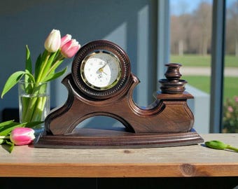 Puede incluir: Un reloj de repisa de madera marrón oscuro con una esfera redonda y ribete dorado. El reloj tiene una base decorativa y curvada y un diseño tallado detallado. El reloj está sobre una superficie de madera.