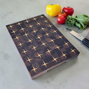 Puede incluir: Una tabla de cortar rectangular de madera de testa con un patrón geométrico marrón oscuro y detalles claros en forma de estrella. La tabla está colocada junto a un pimiento amarillo, dos tomates rojos, un manojo de hierbas verdes y un cuchillo.