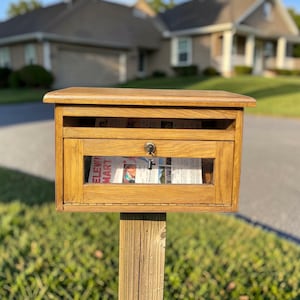 May include: A wooden mailbox with a key lock, mounted on a wooden post. The mailbox has a rectangular shape with a small window on the front, revealing newspapers inside. The wood has a natural, light brown color.