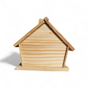 Puede incluir: Una peque&ntilde;a caja de madera con forma de casa. La casa tiene un acabado de madera natural de color marr&oacute;n claro con un tejado a dos aguas y un peque&ntilde;o detalle de chimenea. La caja se asienta sobre una base rectangular y se aprecia la veta de la madera.