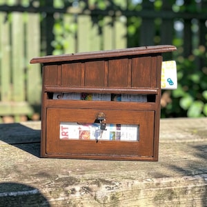 May include: A wooden mailbox with a slanted roof and a lockable door. The mailbox is stained a dark brown color and has a slot for mail and a window to see the contents. Keys are in the lock. Newspapers are visible inside.