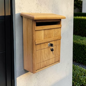 May include: A wall-mounted wooden mailbox featuring a natural light brown wood grain finish, a slanted top lid, a horizontal mail slot, and a front-opening door secured by a silver metal lock with a black key inserted into the keyhole.