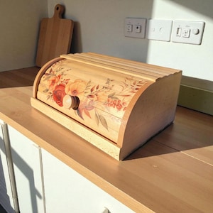 Puede incluir: Panera de madera con tapa enrollable. La panera está decorada con un diseño floral en tonos rojos, naranjas y marrones. Una tabla de cortar de madera está en el fondo. La panera está sobre una encimera de madera.