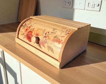 May include: A light-colored wooden bread box with a roll-top lid featuring a floral print of red and orange flowers, leaves, and berries. The bread box has a small round wooden knob on the front and is positioned on a light wood countertop.