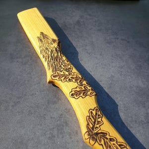 Può includere: Un'intaglio in legno raffigurante un lupo che ulula e foglie di quercia. Il legno di colore chiaro è intagliato in modo intricato con la testa del lupo e dettagliati disegni di foglie di quercia. L'intaglio ha una venatura naturale del legno e una finitura liscia.
