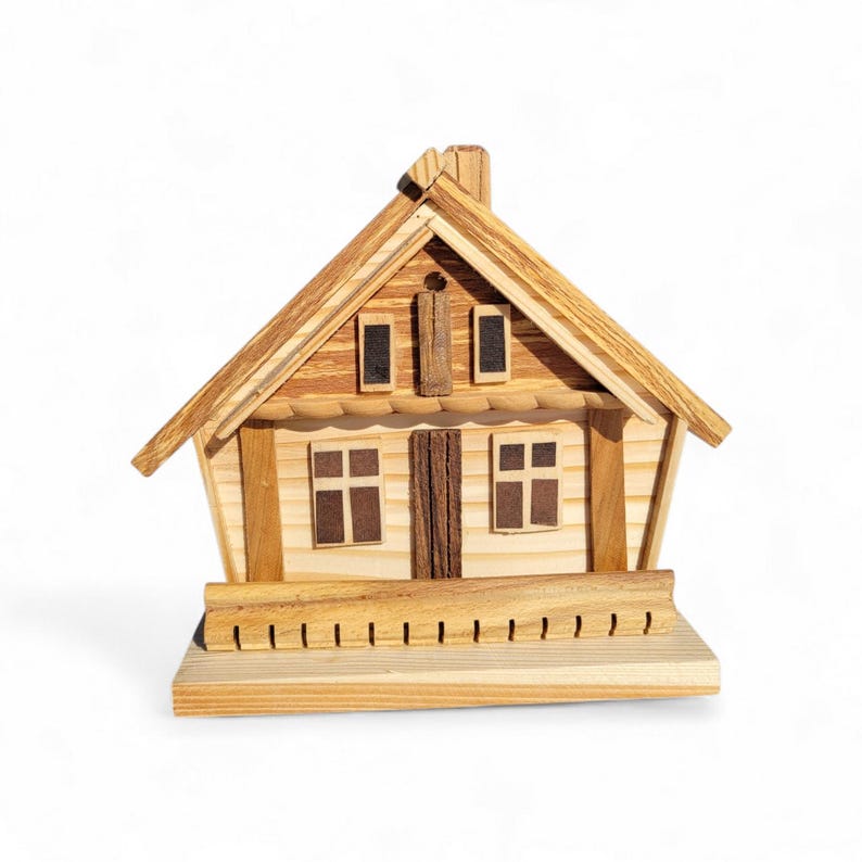 Puede incluir: Una casa de madera en miniatura con tejado a dos aguas, chimenea y peque&ntilde;as ventanas. La casa est&aacute; construida con varias tonalidades de madera clara y oscura, con una base tipo porche. El dise&ntilde;o es detallado y parece hecho a mano.