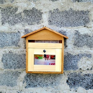 Puede incluir: Un buzón de madera con techo en forma de casa, montado en una pared de piedra gris. El buzón tiene una puerta rectangular con una pequeña ventana que muestra contenido colorido. Una llave cuelga de la cerradura. El buzón es de madera clara.