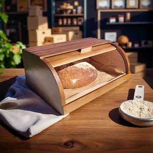 May include: A wooden bread bin with a curved top and a loaf of bread inside. The bin is made of light-coloured wood with a darker wood top. A white cloth and a small bowl of flour are on the wooden table.