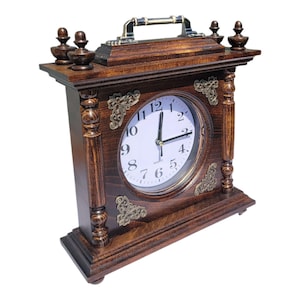 Può includere: Un orologio da mensola in legno marrone scuro con quadrante bianco e numeri neri. L'orologio ha dettagli decorativi in ottone e una maniglia sulla parte superiore. L'orologio è alto circa 25 cm.