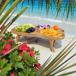 May include: A wooden picnic table with a natural finish, set on a sandy beach. The table features compartments holding green grapes, blue grapes, cheese, chips, and an orange. The table has folding legs and is surrounded by greenery and the ocean.