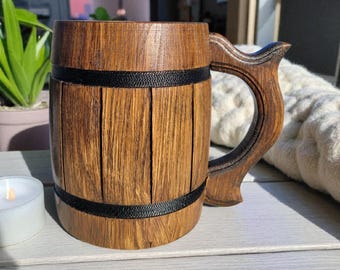May include: A handcrafted wooden beer mug with a decorative handle. The mug is made of vertical wooden planks, bound by two black bands. The handle is intricately carved. The mug is a rich brown color.
