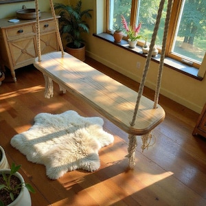 Puede incluir: Un columpio de madera suspendido por cuerdas. El asiento del columpio es rectangular y de madera clara. Una alfombra cremosa y esponjosa está sobre el suelo de madera debajo del columpio. La habitación tiene una ventana con luz natural.