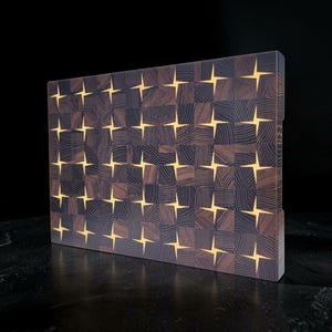 Pode incluir: Uma tábua de corte retangular de madeira de topo com uma grade de blocos de madeira castanho-escuro em padrão xadrez, separados por incrustações de resina clara em forma de estrela que criam um design geométrico repetitivo.