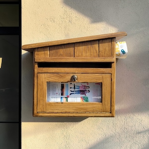 May include: A wooden mailbox with a slanted roof is mounted on a textured wall. The mailbox has a glass window displaying printed materials and a small lock. A roll of paper with a yellow and blue pattern is tucked into the side.